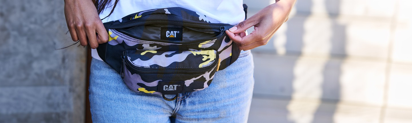 Person opening a camouflage CAT waist bag while wearing jeans and a white shirt.