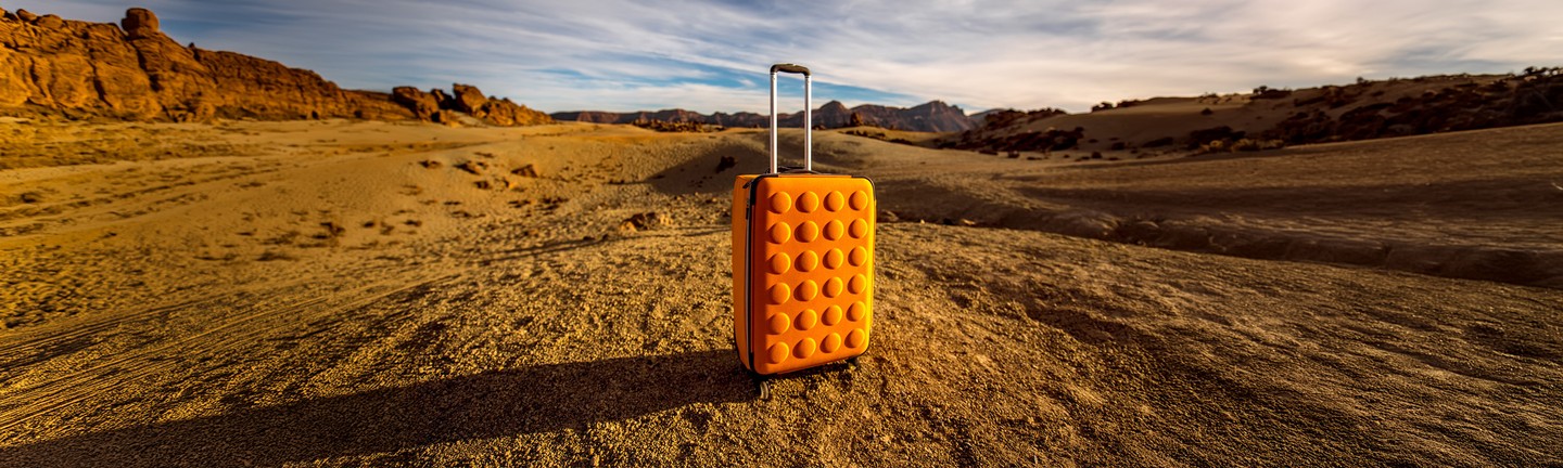 Orange suitcase standing alone in a vast desert landscape with rocky mountains in the background.