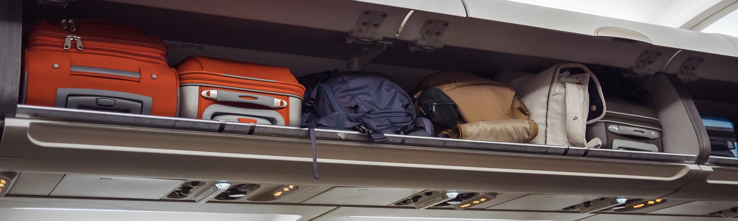 Overhead compartment in an airplane filled with various types of carry-on luggage, including orange suitcases and backpacks