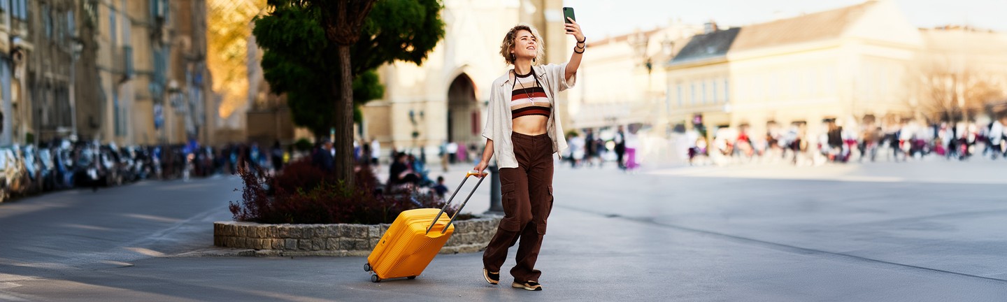 Young woman taking a selfie while pulling a yellow rolling suitcase on a busy city street.