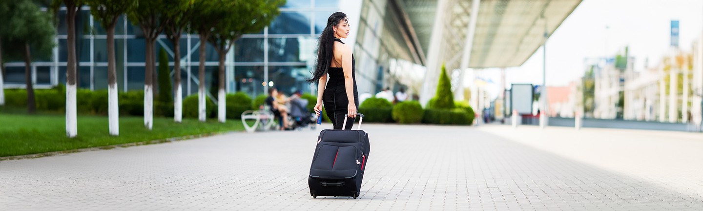 Woman in a black dress walking with a black rolling suitcase on a city street, with modern buildings in the background.