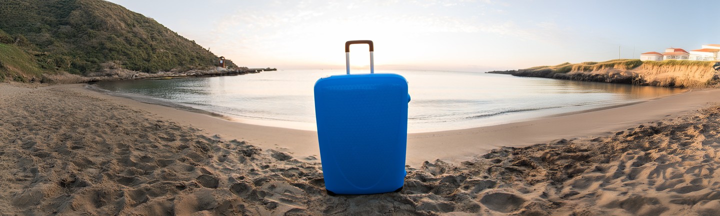 Blue suitcase standing on a sandy beach with a calm sea and green hills in the background.